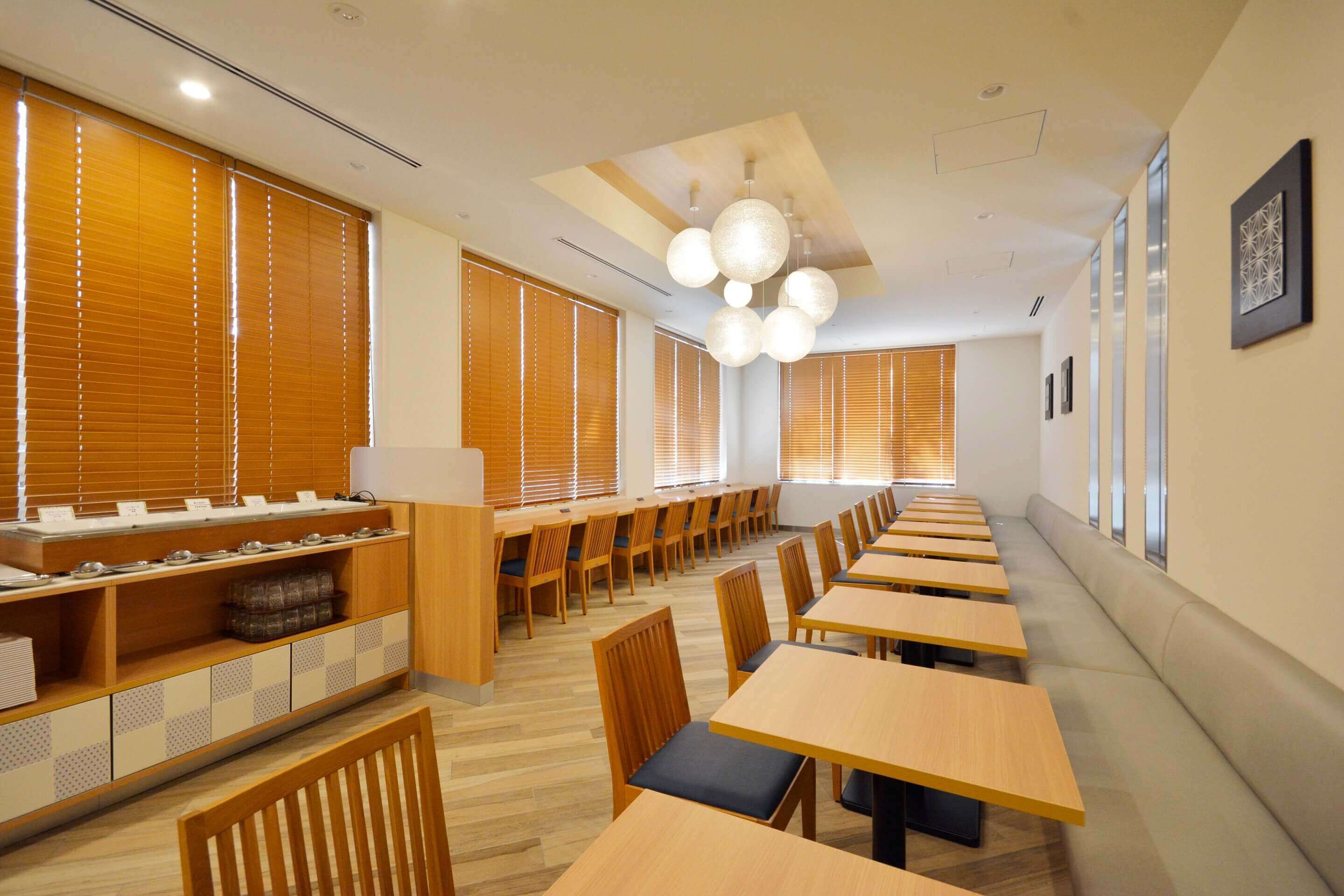 Un'area pranzo moderna con lunghe tavole e sedie in legno, una postazione di servizio centrale e ampie finestre con tende.