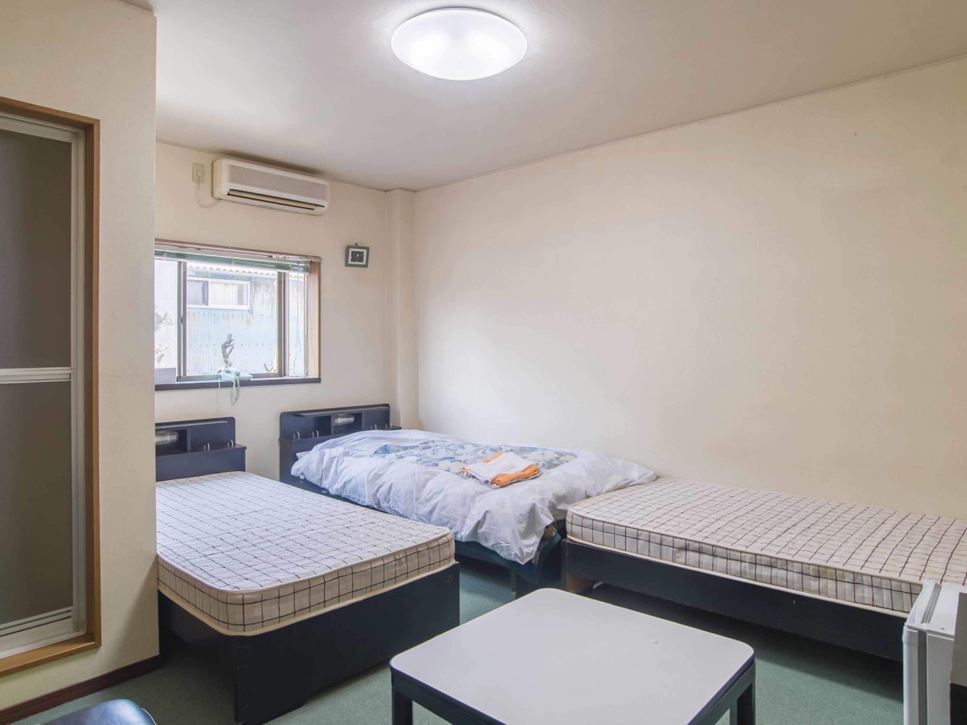 A small, simple room with two single beds, a small table, and a window with a view of the outside.