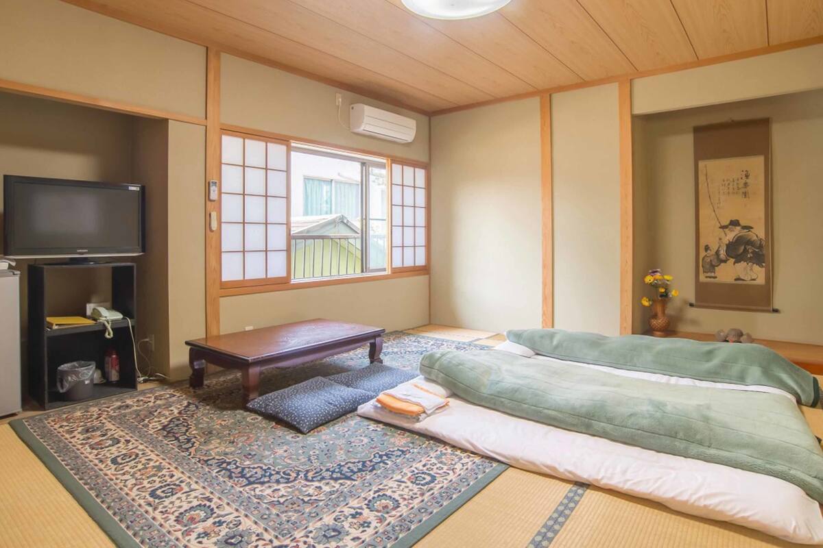 Une pièce de style japonais traditionnel, avec un sol recouvert de tatamis, une basse table en bois, un téléviseur et une porte coulissante donnant sur l’extérieur.