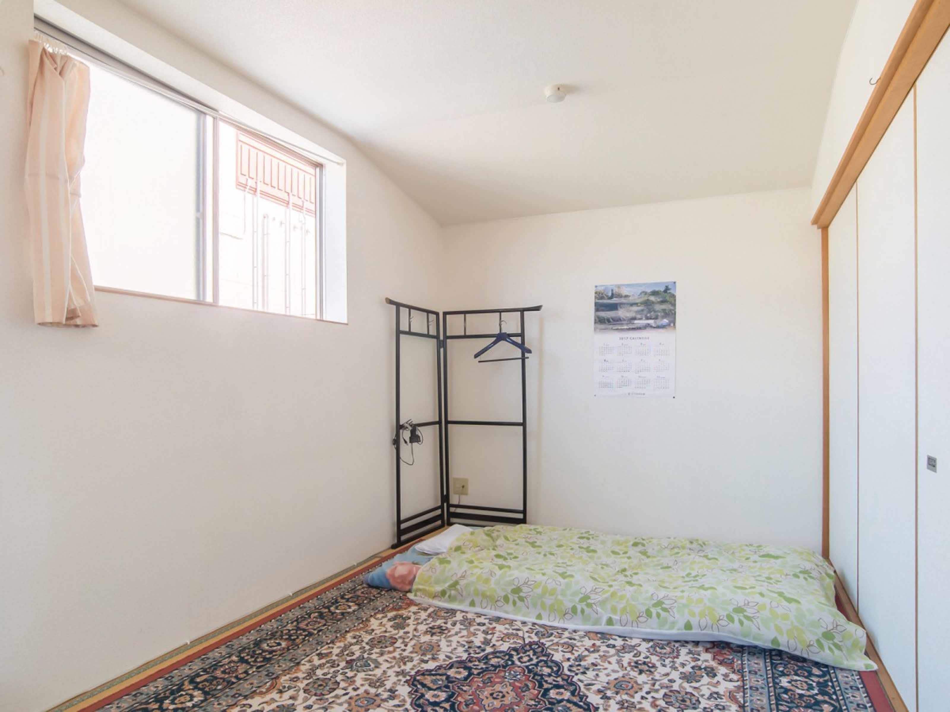 A small, sparsely furnished room with a single bed, a window with curtains, a radiator, and a calendar on the wall.