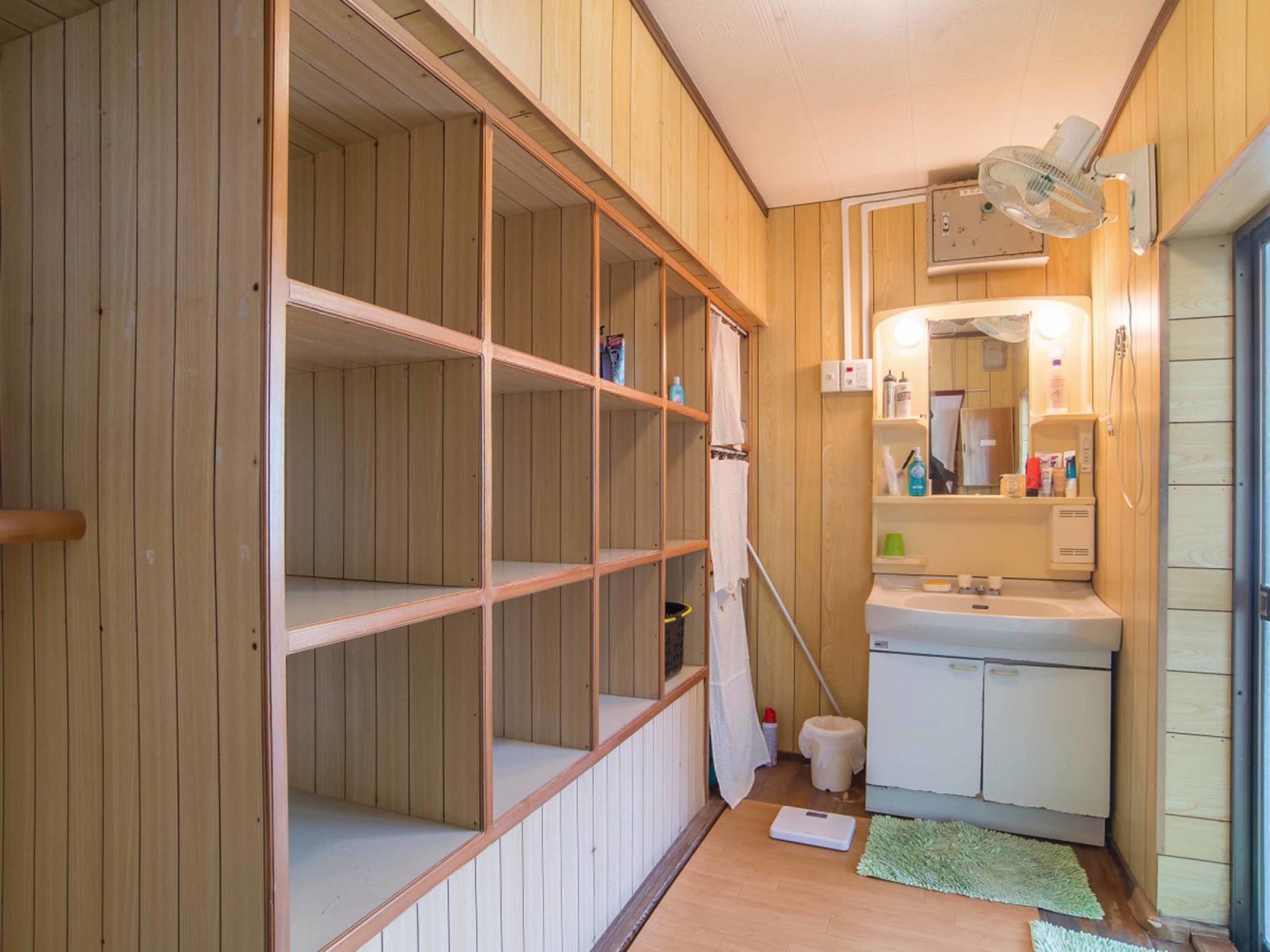 Un baño pequeño y funcional con paredes de madera, un lavabo blanco y un espejo. Hay estantes sobre el lavabo y un taburete a su lado.