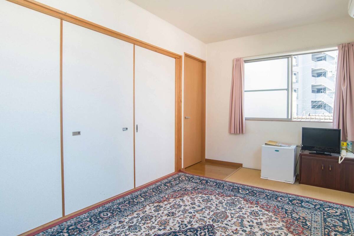 Une chambre dotée d’une grande armoire, d’un petit réfrigérateur, d’une télévision et d’une fenêtre avec des rideaux.