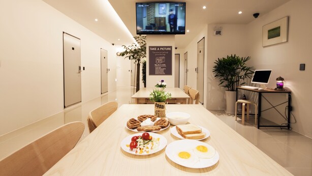 A modern hotel room with a dining table set with waffles, eggs, and toast. A flat-screen TV is mounted on the wall, and there is a sign encouraging guests to take pictures. - Seoul Grand Hostel Ewha Univ. (Seoul)