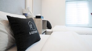 A hotel room with a bed, a nightstand, a mirror, a window with blinds, and a pillow with the text "K-GUESTHOUSE". - Seoul Grand Hostel Ewha Univ. (Seoul)