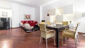 Un salon moderne avec un canapé rouge, une table à manger entourée de chaises en osier et, au fond, un coin cuisine.