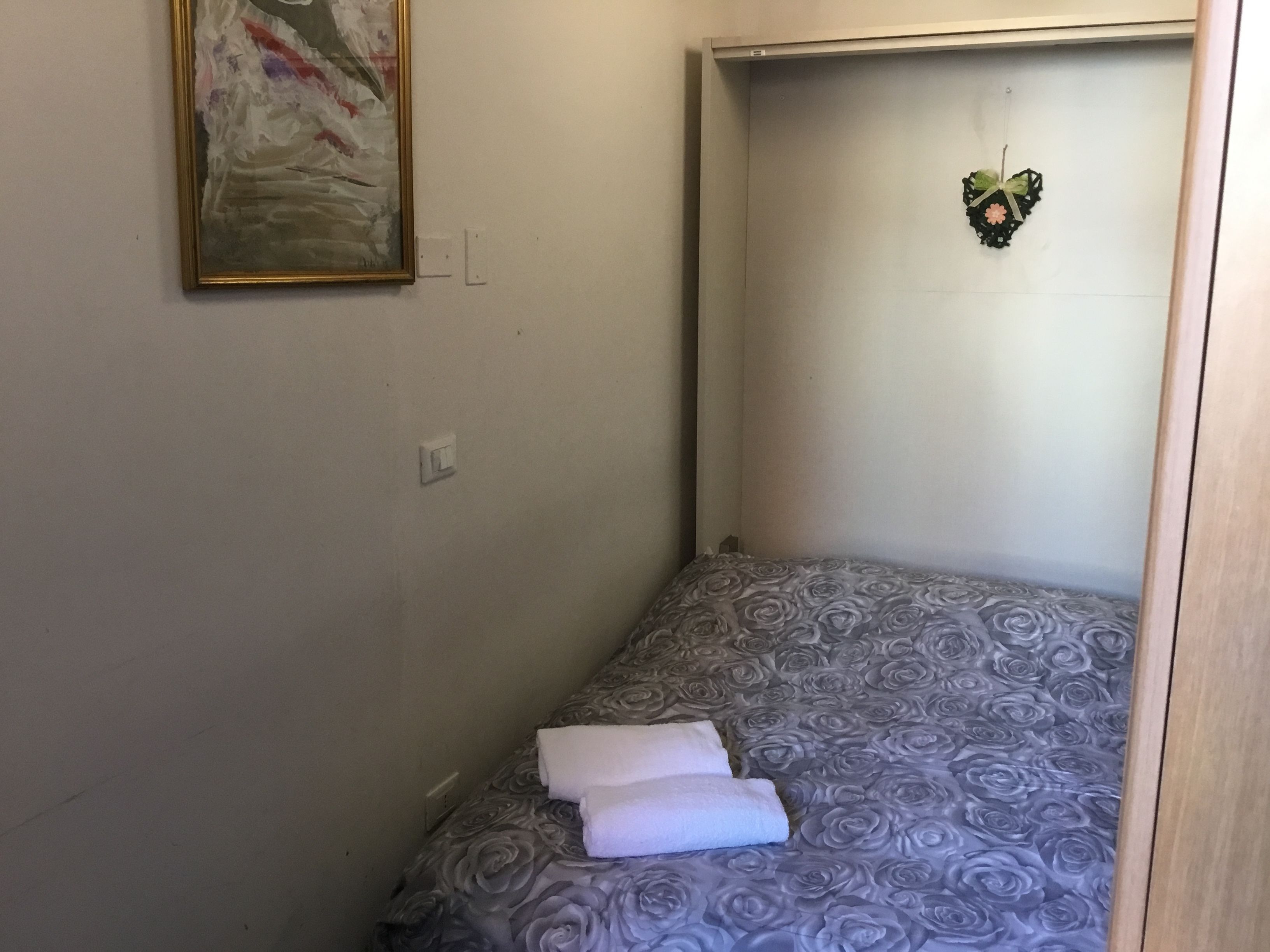 A small, single-bed room with a grey floral bedspread, a painting on the wall, and a heart-shaped decoration.