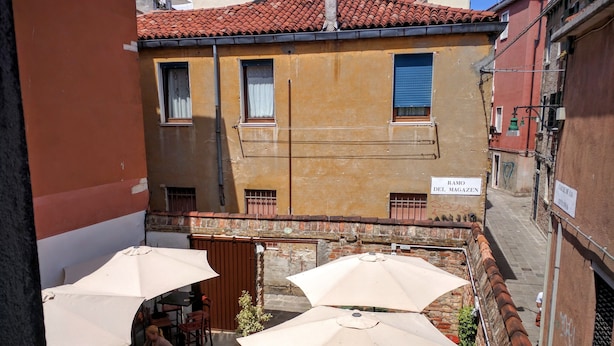 Un área de estar al aire libre con mesas y sombrillas, un muro de ladrillo y un edificio con un letrero que dice "RAMO DEL MAGAZEN".