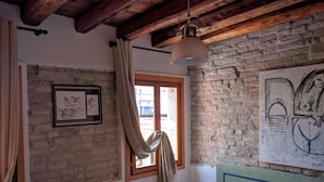 Una habitación rústica con paredes de ladrillo visto, techo de madera, cama con cabecera verde, un cuadro enmarcado en la pared y una ventana con cortinas.