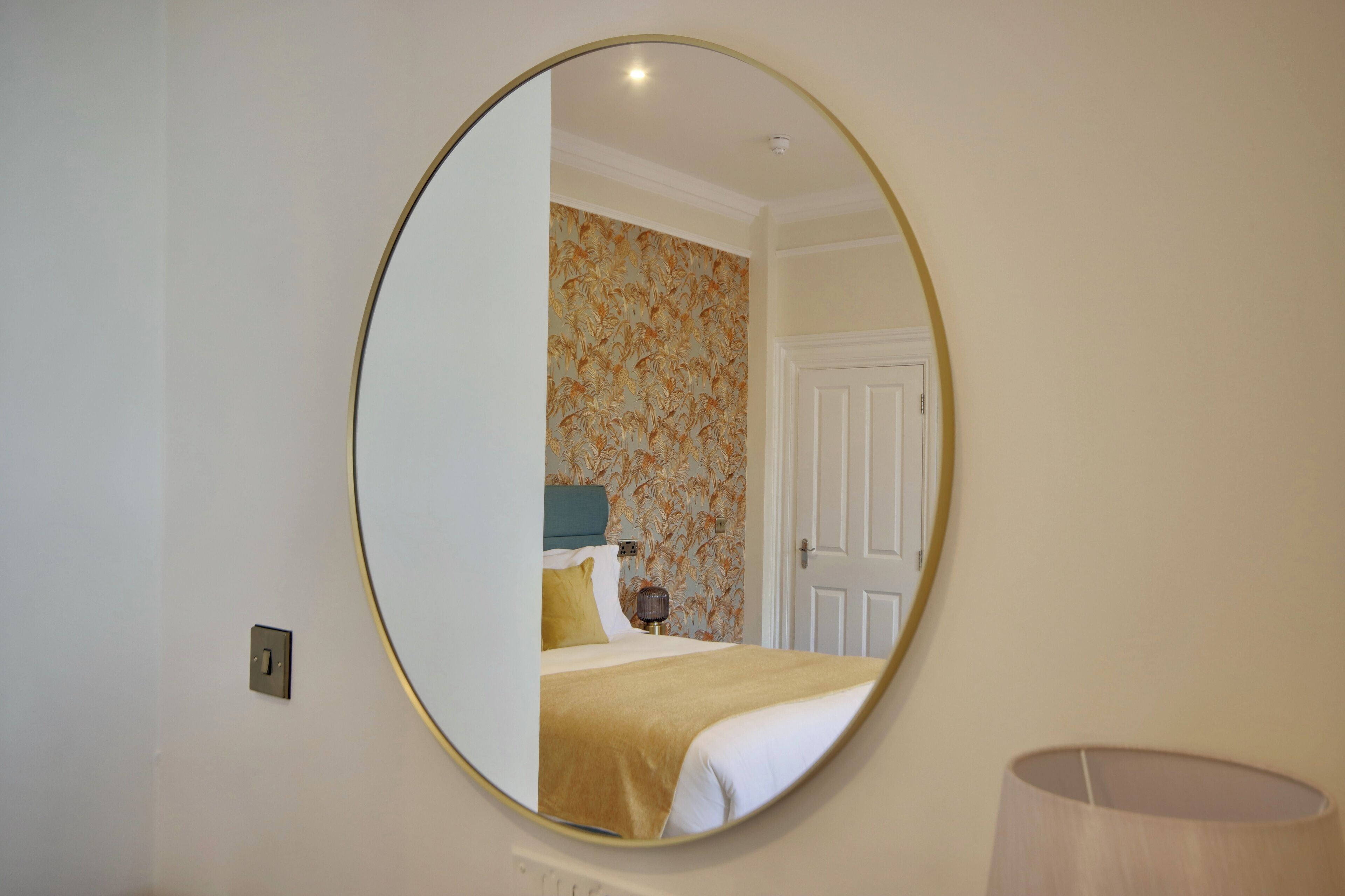 Een kamer met een grote ronde spiegel aan de muur, een bed met hoofdeinde, een nachtkastje en een lamp.