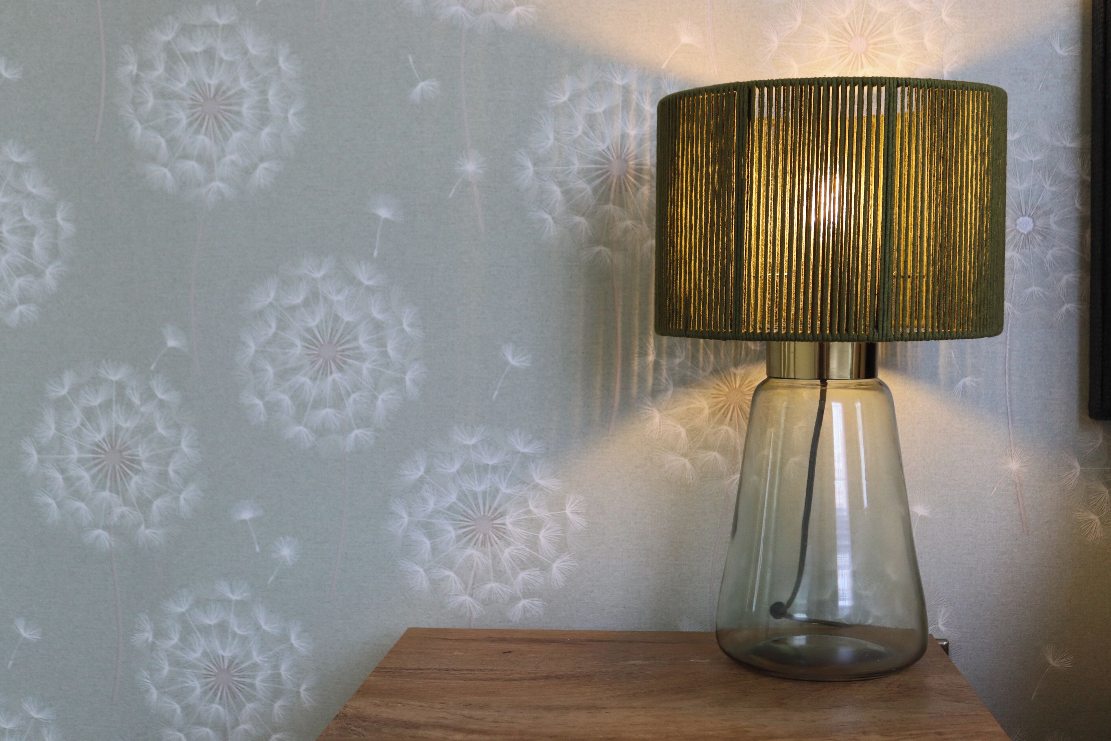 A lamp with a textured shade on a wooden surface against a wall with a dandelion pattern.