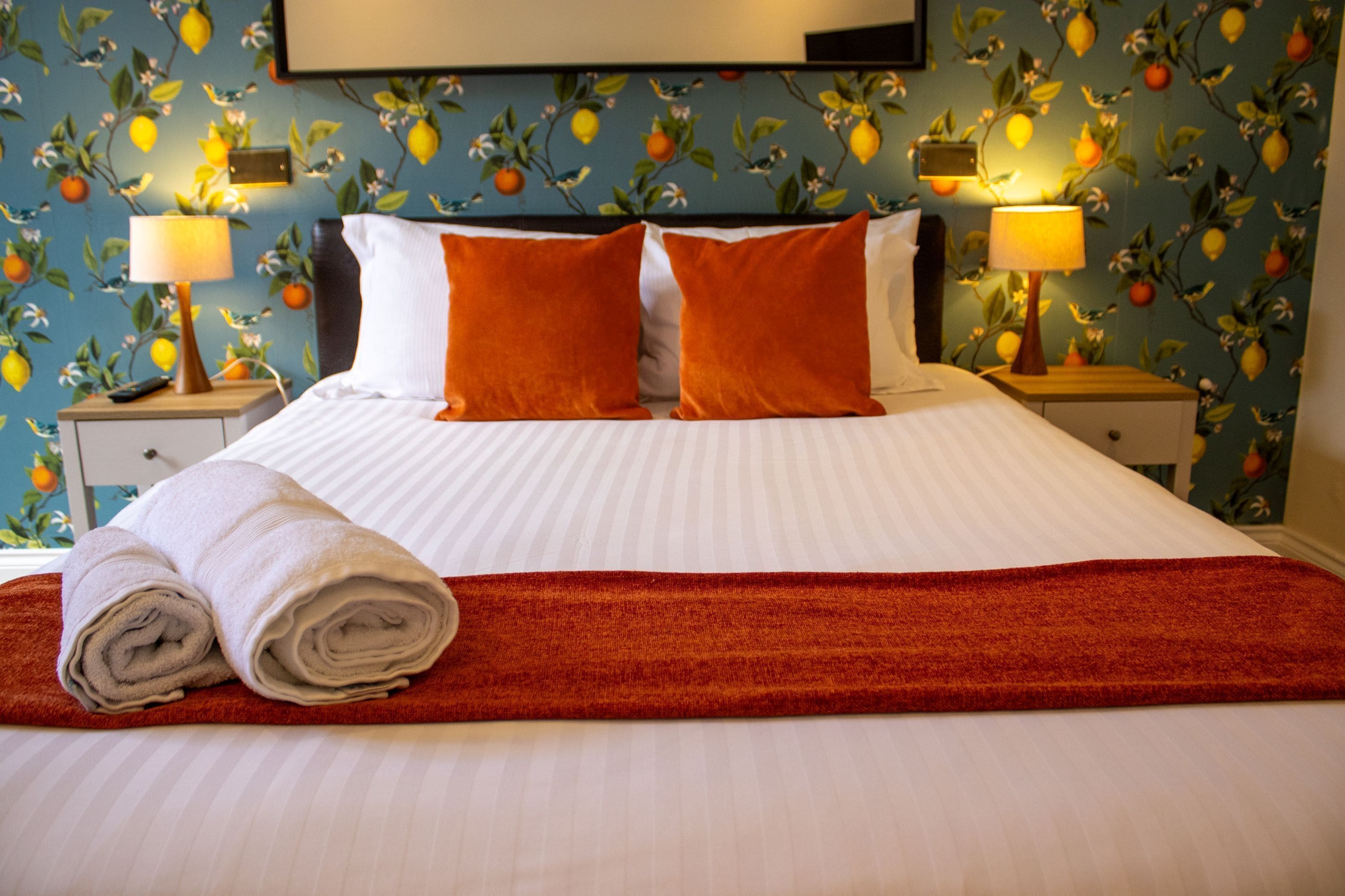 A hotel room with a bed, two nightstands, a red throw blanket, and a wall with a fruit pattern.