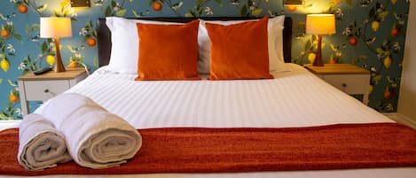 A hotel room with a bed, two nightstands, a red throw blanket, and a wall with a fruit pattern.