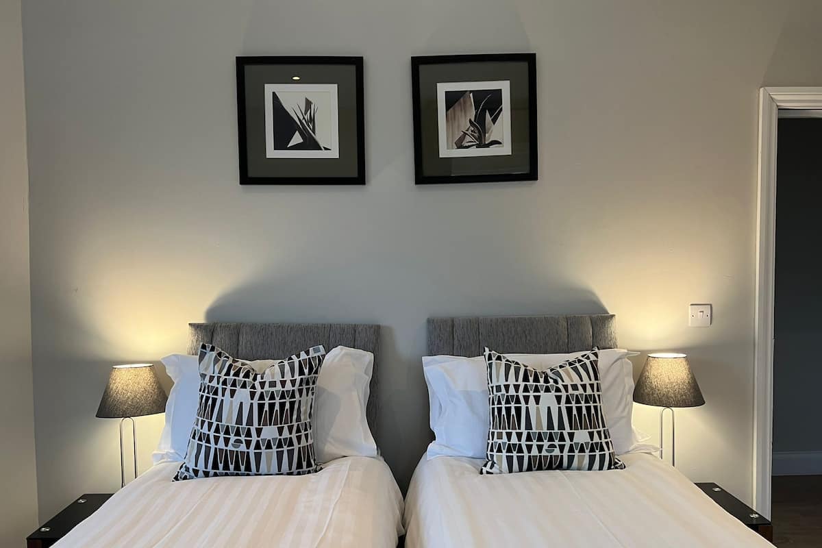A double bed with striped bedding and two pillows, flanked by nightstands with lamps, in a room with a plain wall and two framed pictures above the bed.