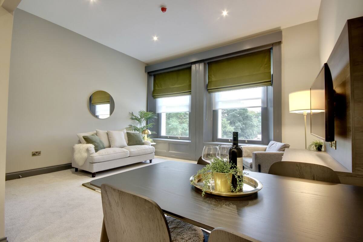 A modern living room with a dining table, sofas, and large windows with green blinds.