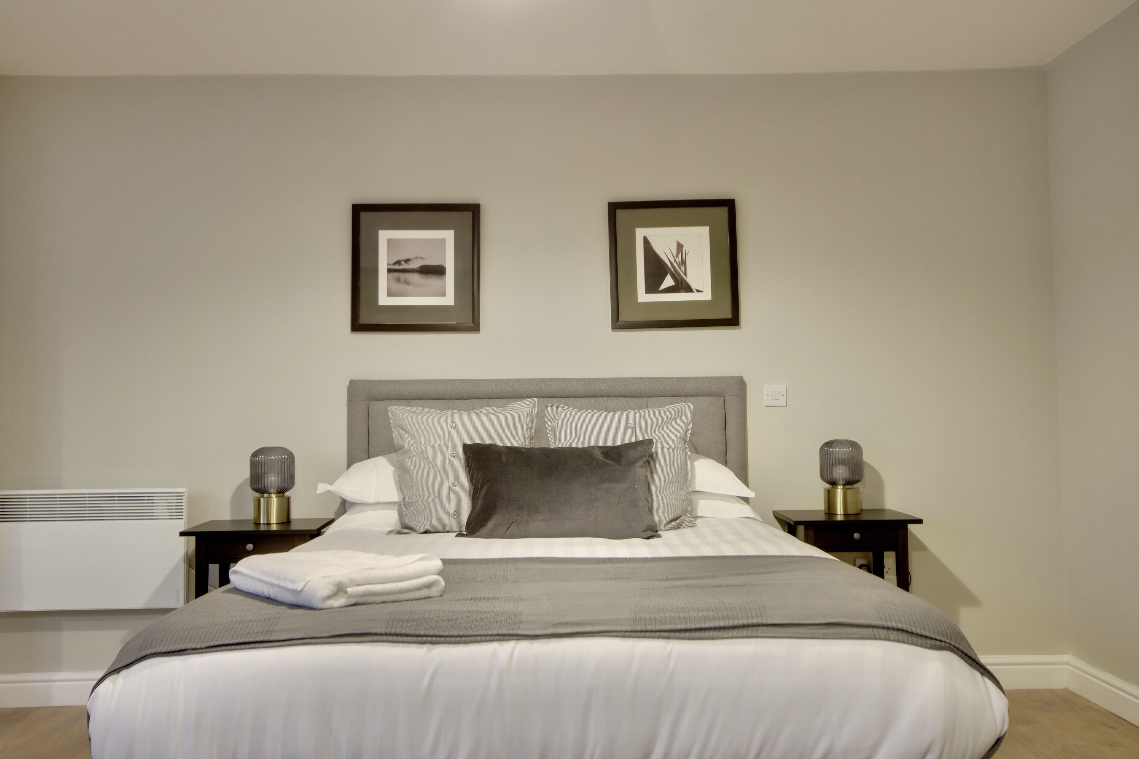 A neatly made bed with a grey blanket, two bedside lamps, and two framed pictures on the wall.