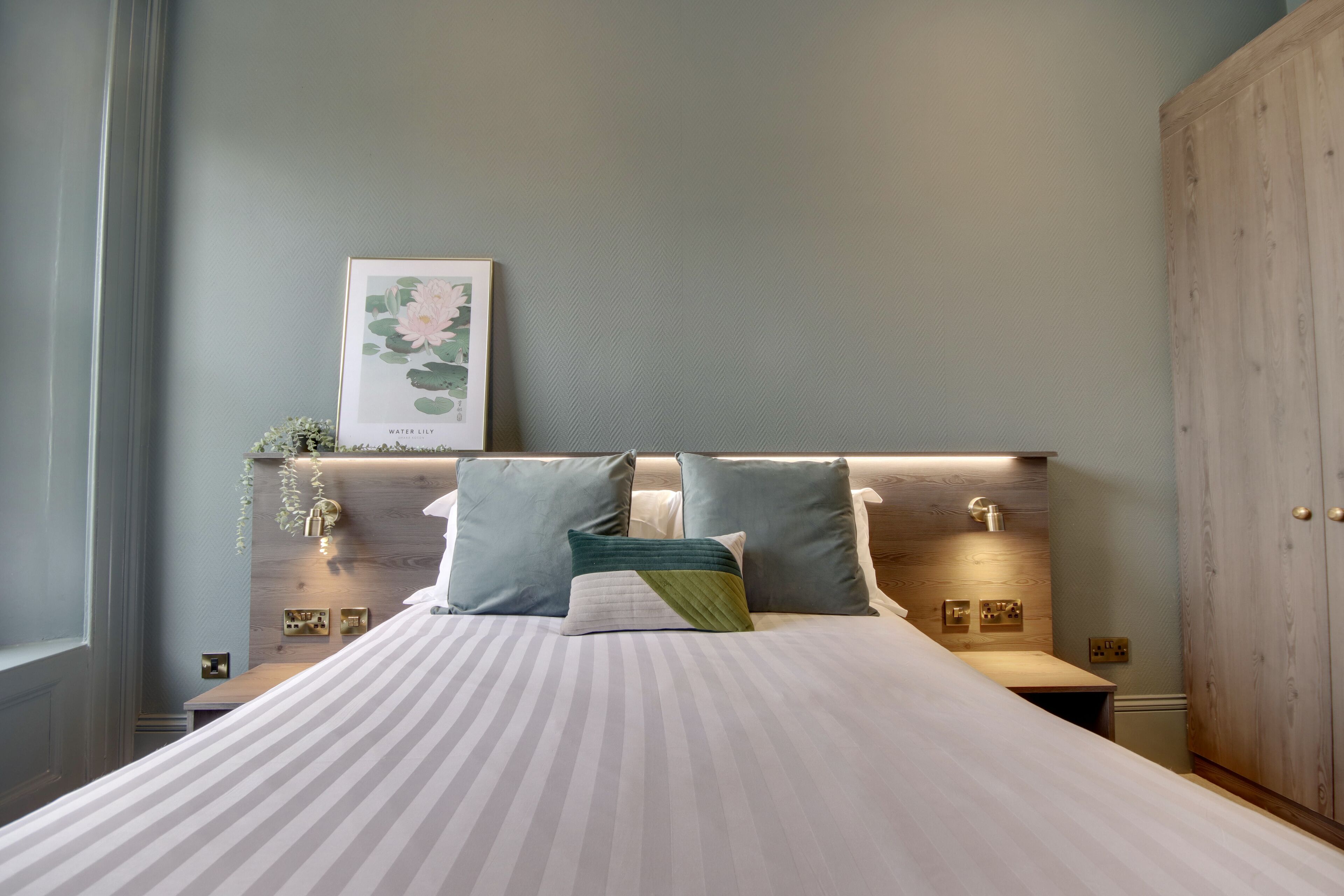 A neatly arranged bedroom with a large bed, two bedside tables, a wall-mounted shelf with decorative items, and a wardrobe.