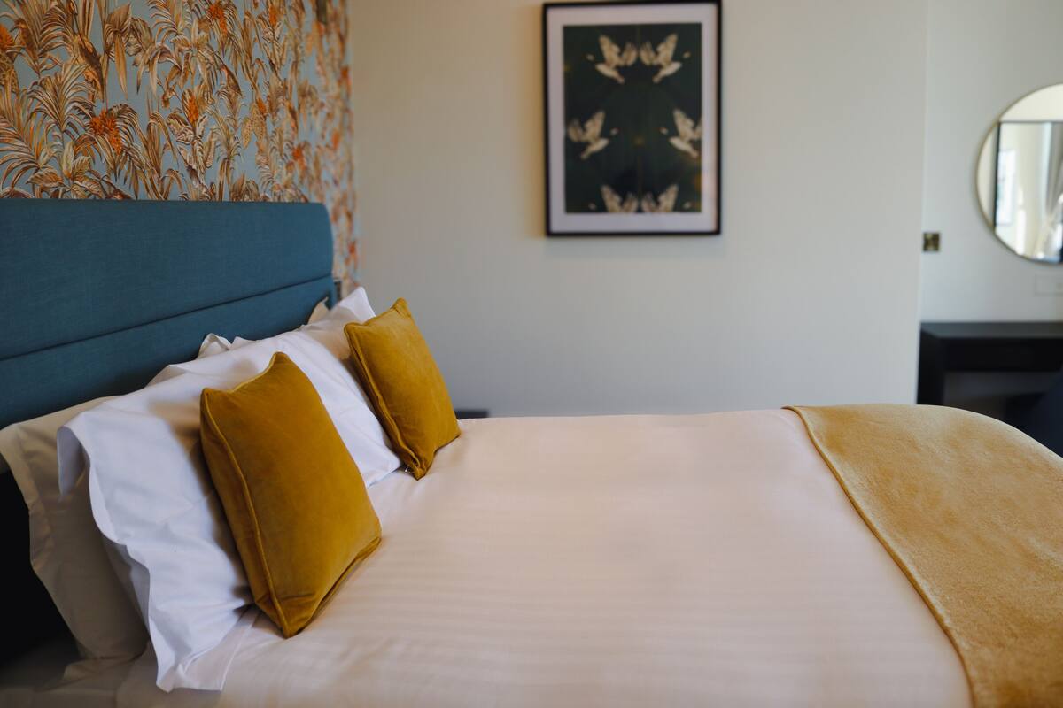 A neatly made bed with white and mustard pillows, a blue headboard, and a framed butterfly artwork on the wall.