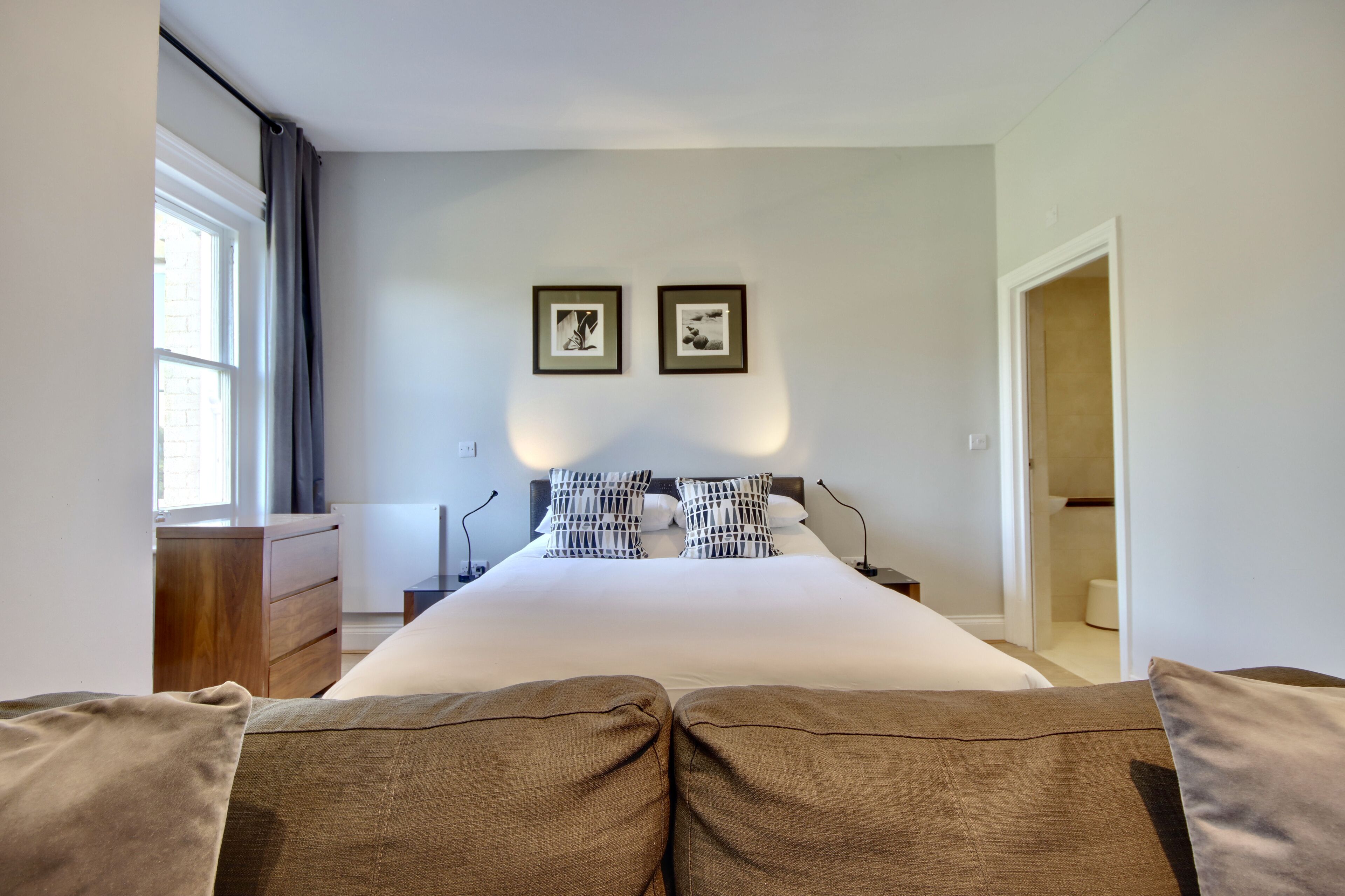 A bedroom with a large bed, a wooden dresser, two framed pictures on the wall, and a door leading to another room.