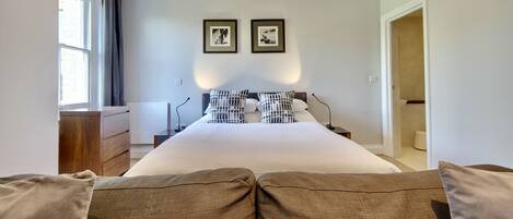 A bedroom with a large bed, a wooden dresser, two framed pictures on the wall, and a door leading to another room.