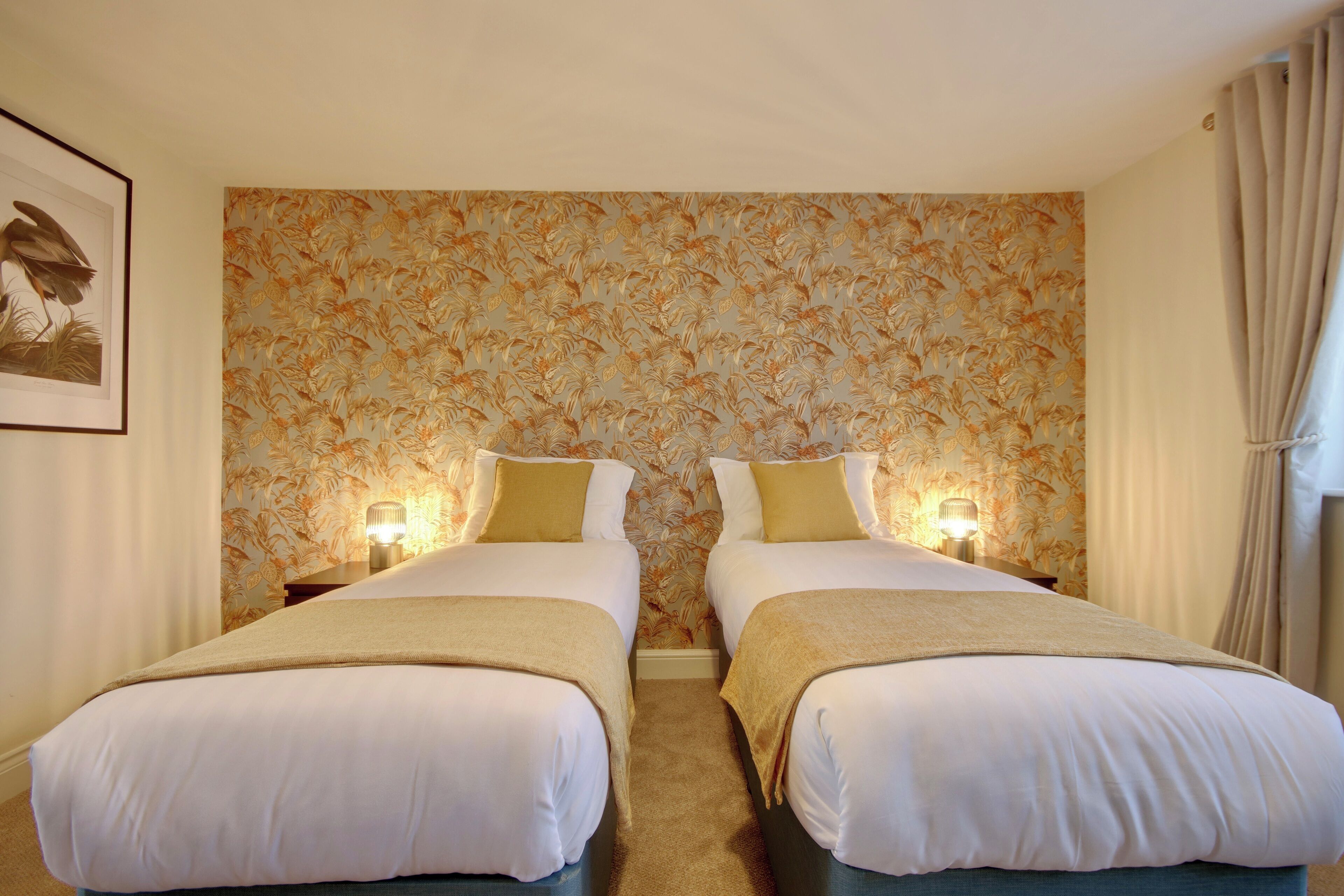 Una camera d'albergo con due letti, biancheria beige, carta da parati floreale e abat-jour.