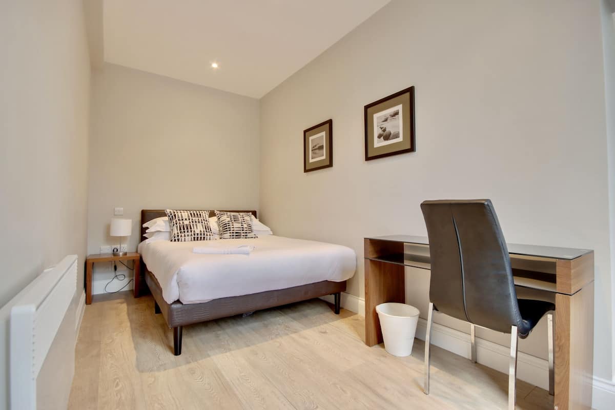A bedroom with a bed, bedside table, lamp, desk, chair, and two framed pictures on the wall.