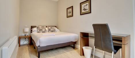 A bedroom with a bed, bedside table, lamp, desk, chair, and two framed pictures on the wall.