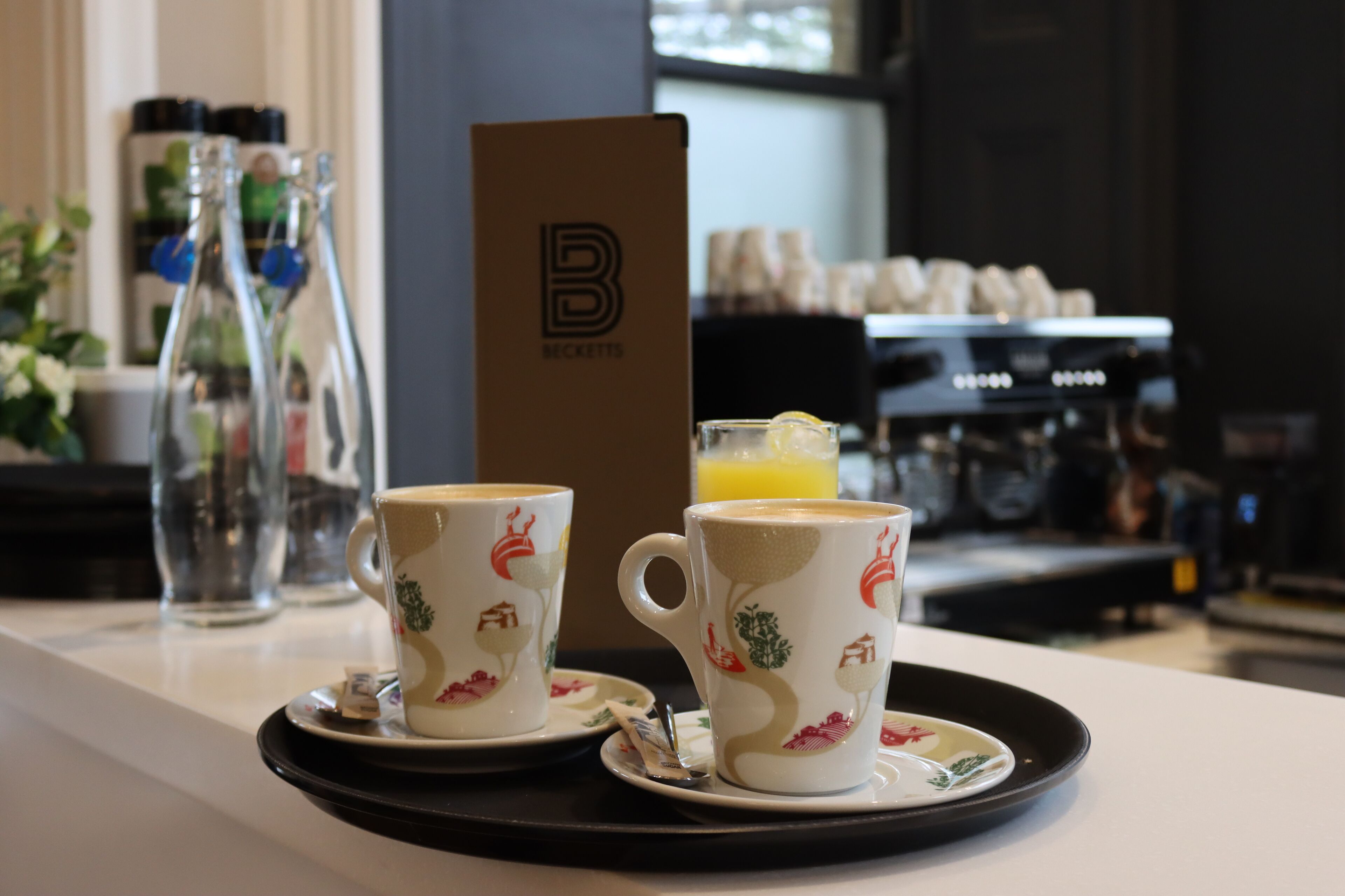 Two cups of coffee on a tray with a glass of orange juice, a coffee machine, and a sign with a logo in the background.
