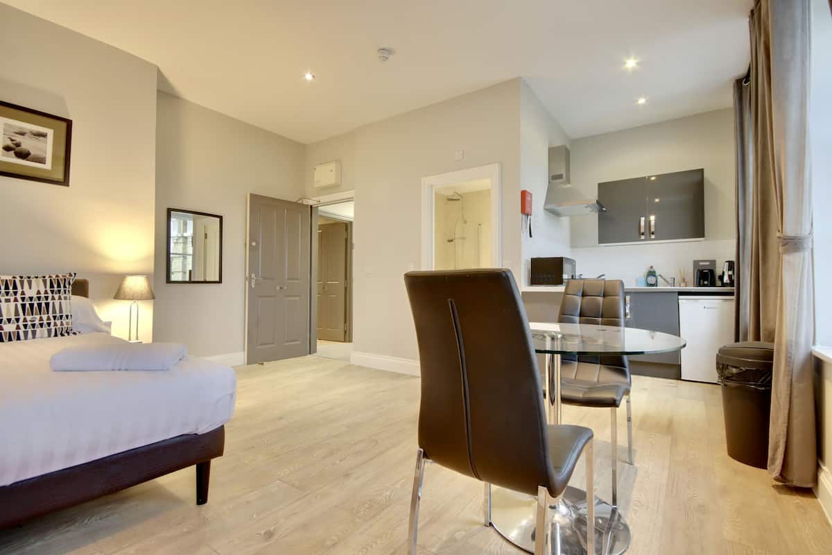 A modern hotel room with a bed, a glass dining table, a chair, a small kitchen area, and a bathroom visible through an open door.
