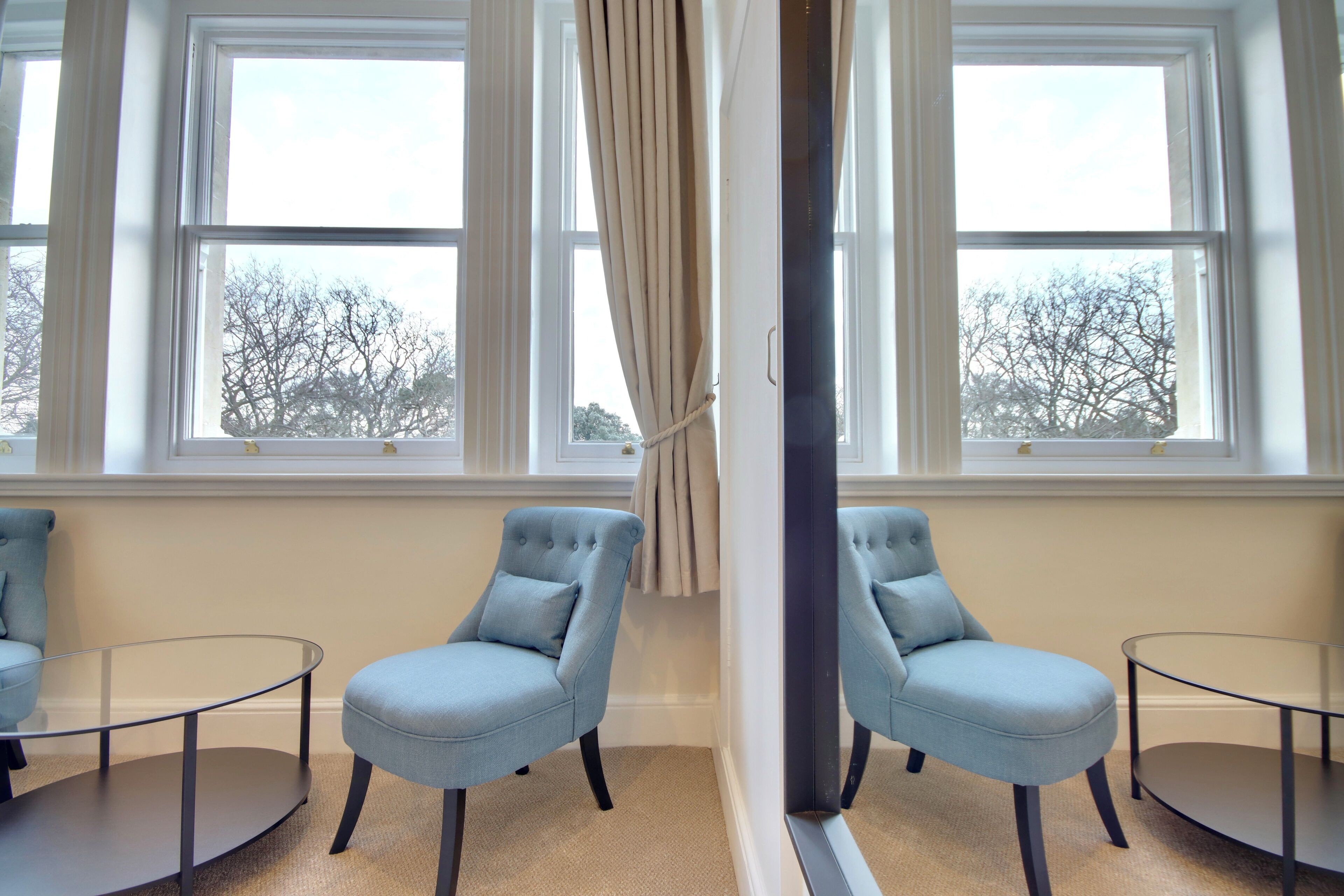 Ein Zimmer mit zwei hellblauen Sesseln, einem Glastisch und einem großen Fenster mit Vorhängen.