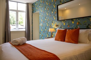 Una habitación de hotel con cama, ventana con cortinas, pared con diseño de frutas y mesita de noche con lámpara.