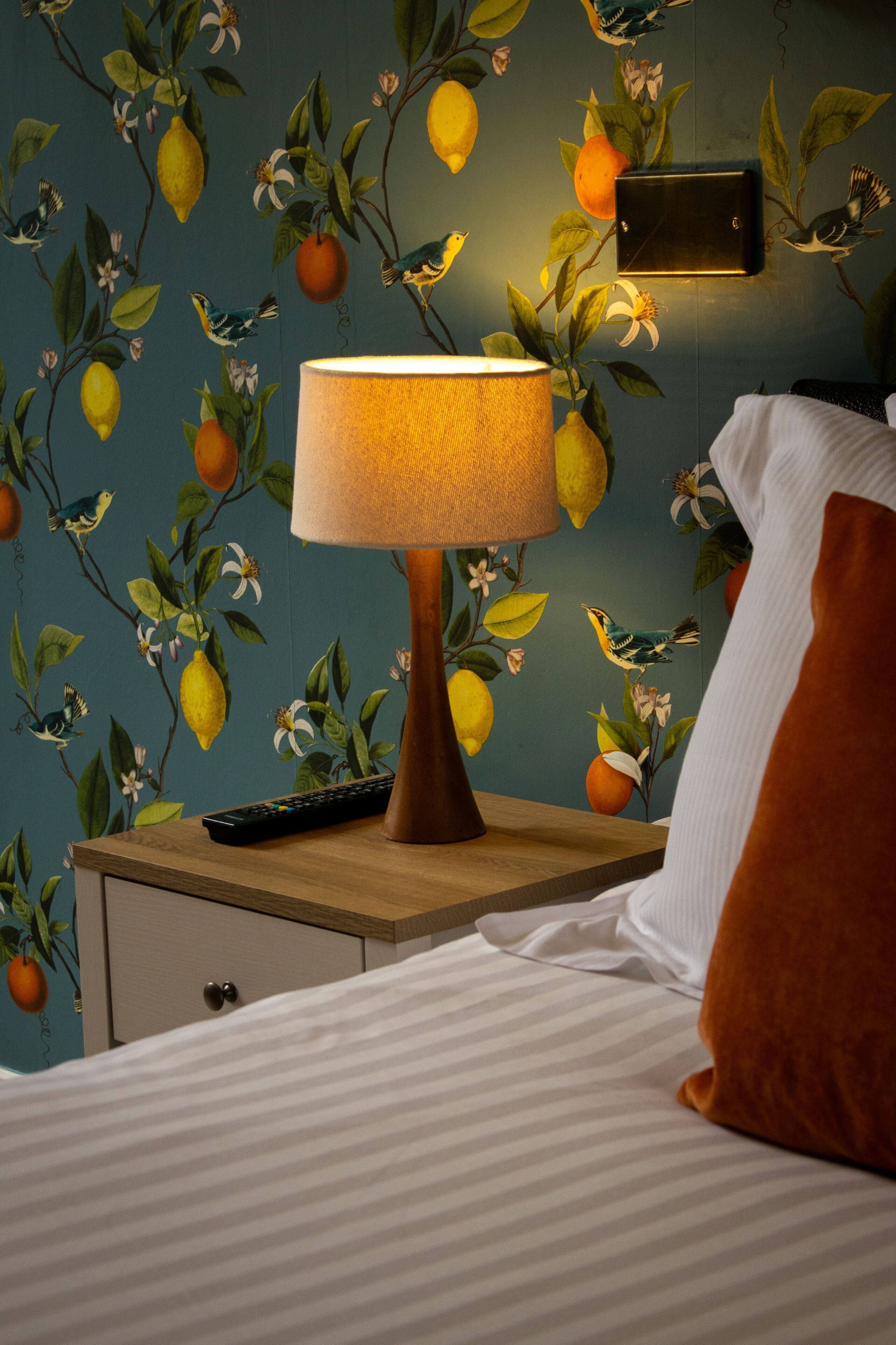 Un dormitorio con cama, mesita de noche, lámpara, control remoto y papel tapiz con motivos de frutas y pájaros.