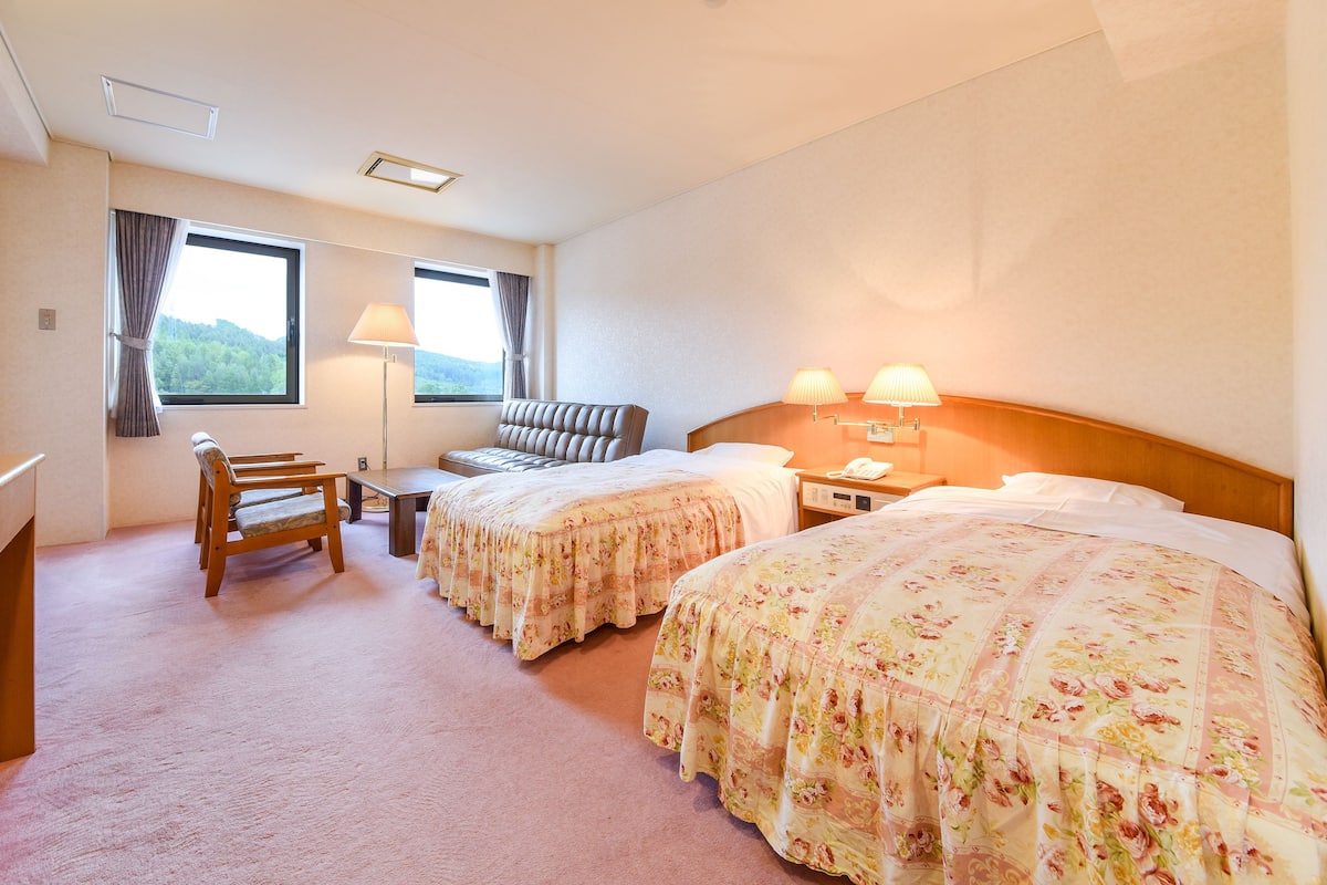 Habitación de hotel con dos camas, un escritorio, una silla, una ventana con cortinas y una lámpara.