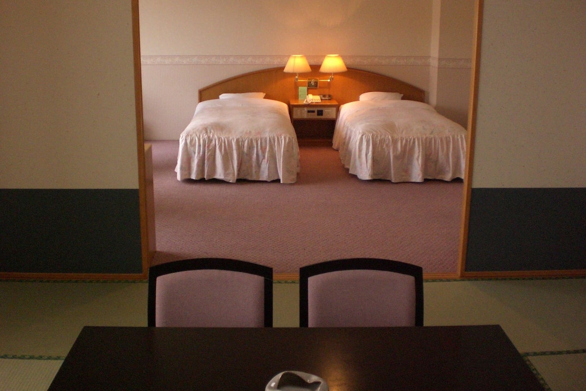 Una habitación de hotel con dos camas individuales, una mesita de noche con lámparas y una pequeña mesa con sillas al frente.