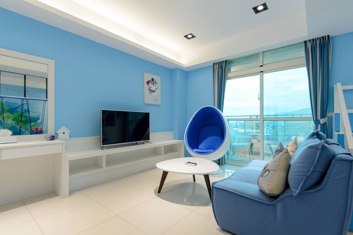 Uma sala de estar moderna com um sofá azul, uma mesa de centro redonda, uma TV de tela plana e uma varanda com vista.