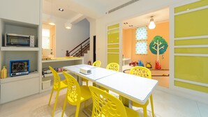 Una zona de menjador àmplia i lluminosa, amb cadires grogues i un taulell blanc, una escala i un mural de paret colorit.