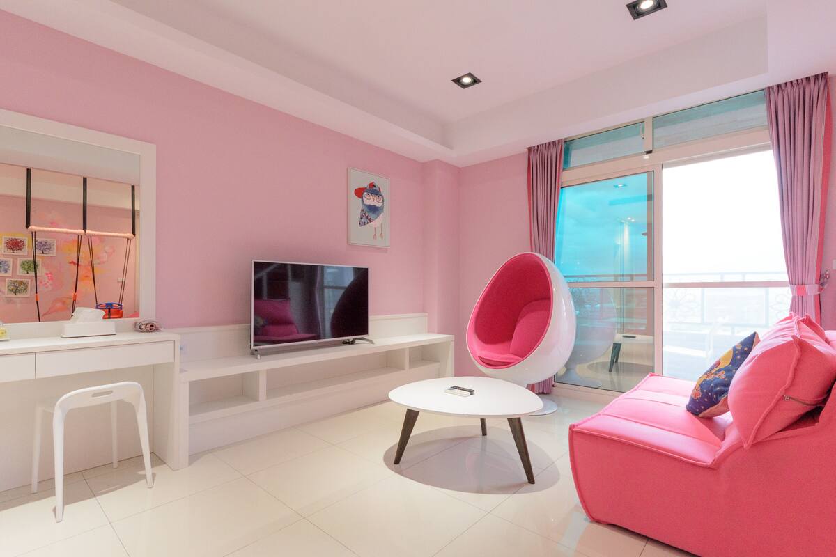 Uma sala de estar em tons de rosa com uma televisão de tela plana, uma escrivaninha branca, uma cadeira branca, uma mesa de centro redonda branca e um sofá rosa com almofadas.