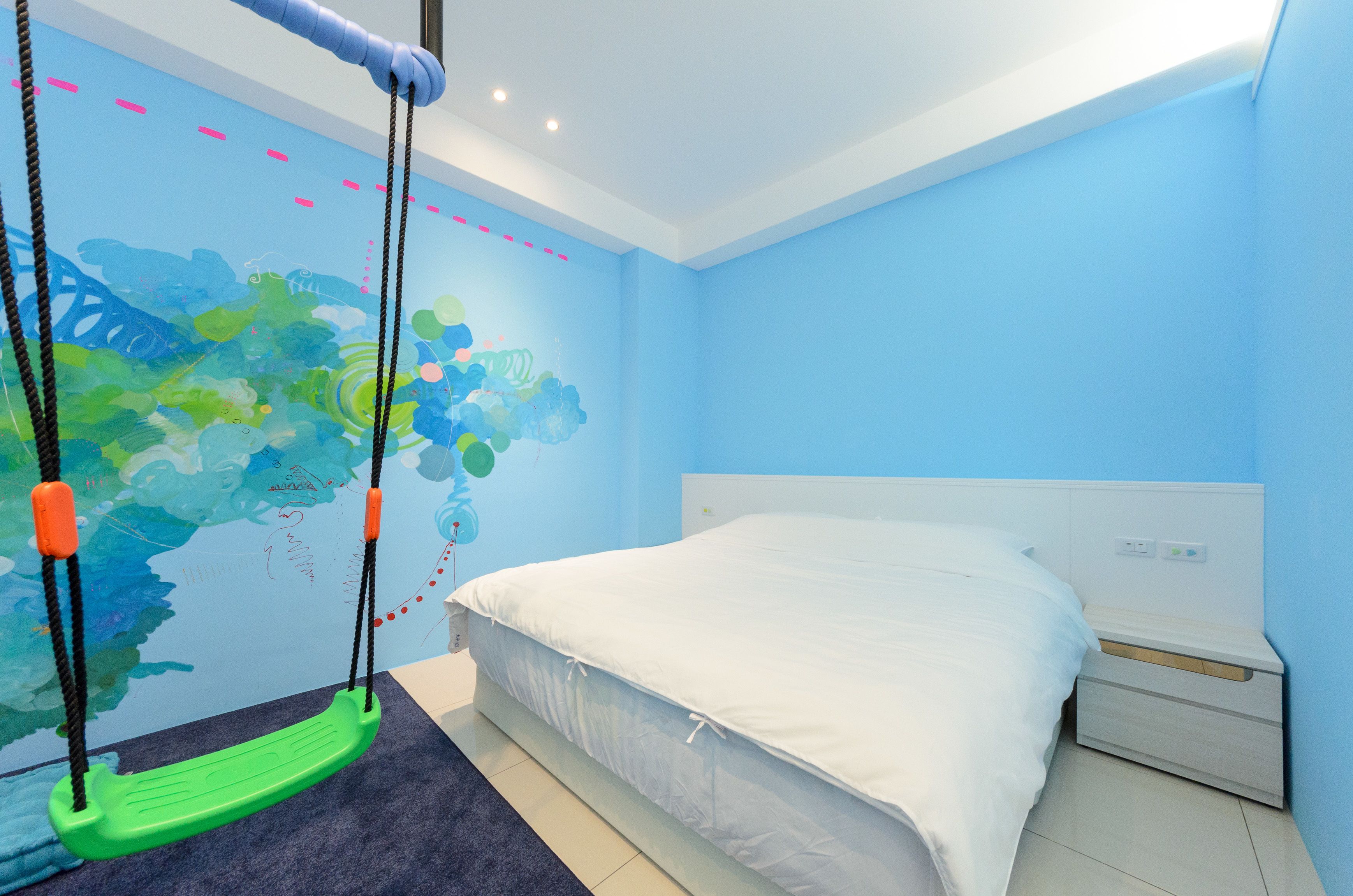A bedroom with a bed, a nightstand, a green swing, and a wall mural.
