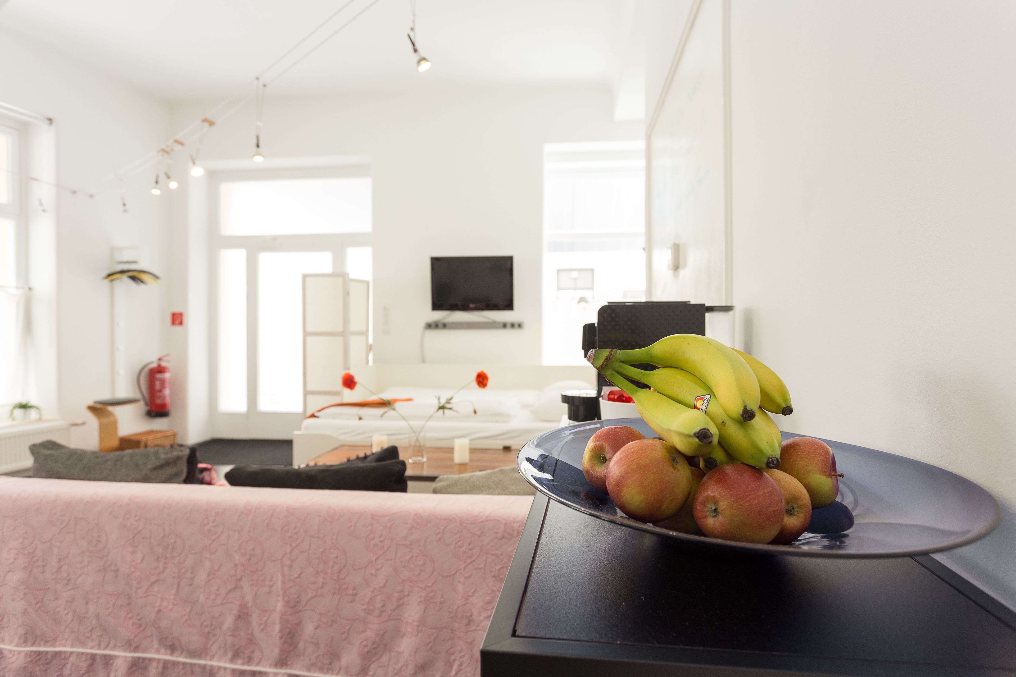 Ein modernes Wohnzimmer mit Essbereich, mit Obstschale auf einem Tisch, einer Couch und einem Fernseher im Hintergrund.