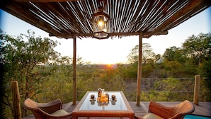 A wooden deck with wicker furniture and a glass table, overlooking a pool and a scenic view. - The Elephant Camp (Victoria Falls)