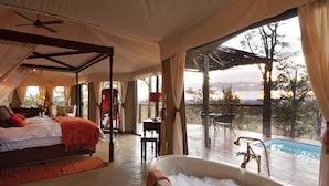 A spacious bedroom with a large bed, a sitting area with a table and chairs, and a view of a swimming pool. - The Elephant Camp (Victoria Falls)