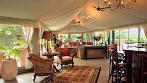 A spacious tented room with a bar area, wicker furniture, and large windows offering a view of the outdoors. - The Elephant Camp (Victoria Falls)