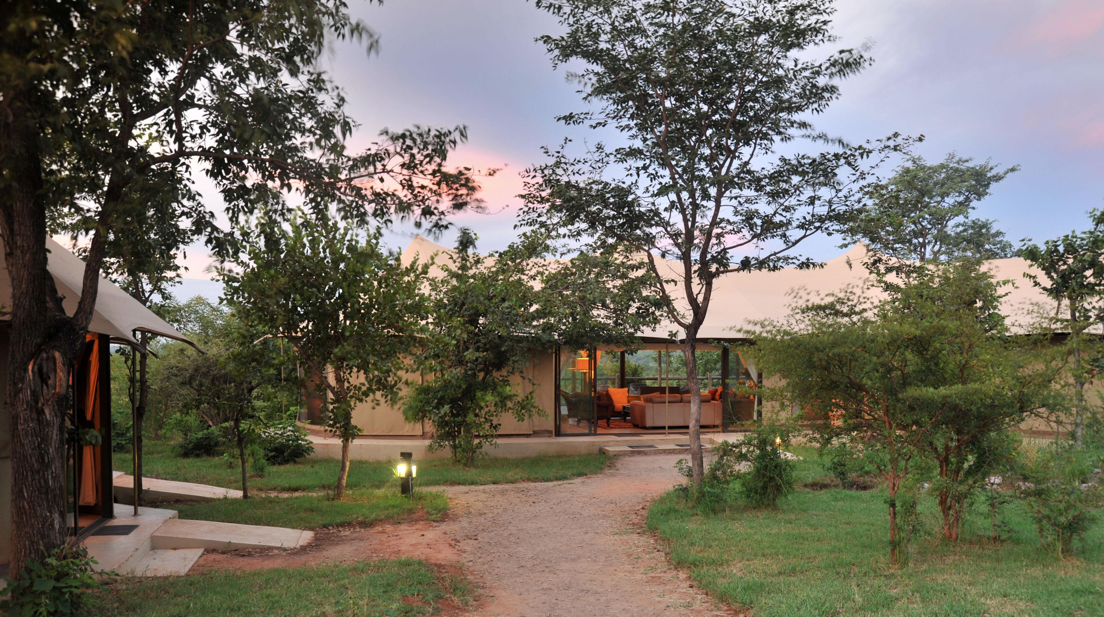 A modern outdoor lounge area with a glass enclosure, surrounded by trees and a dirt path leading to a building with a large glass window.