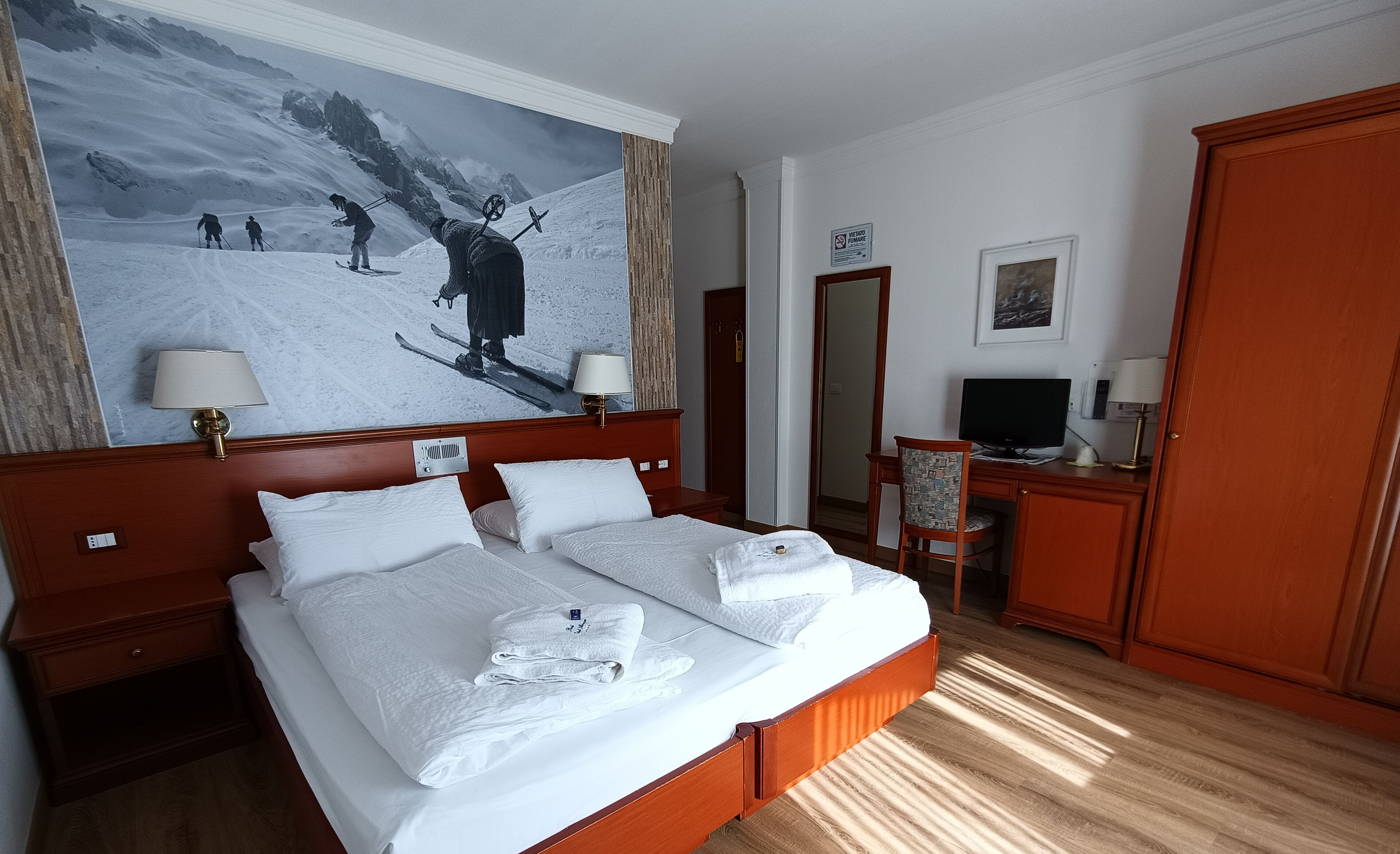 A hotel room with a large ski-themed painting, a bed with white linens, a desk with a computer, and a wardrobe.
