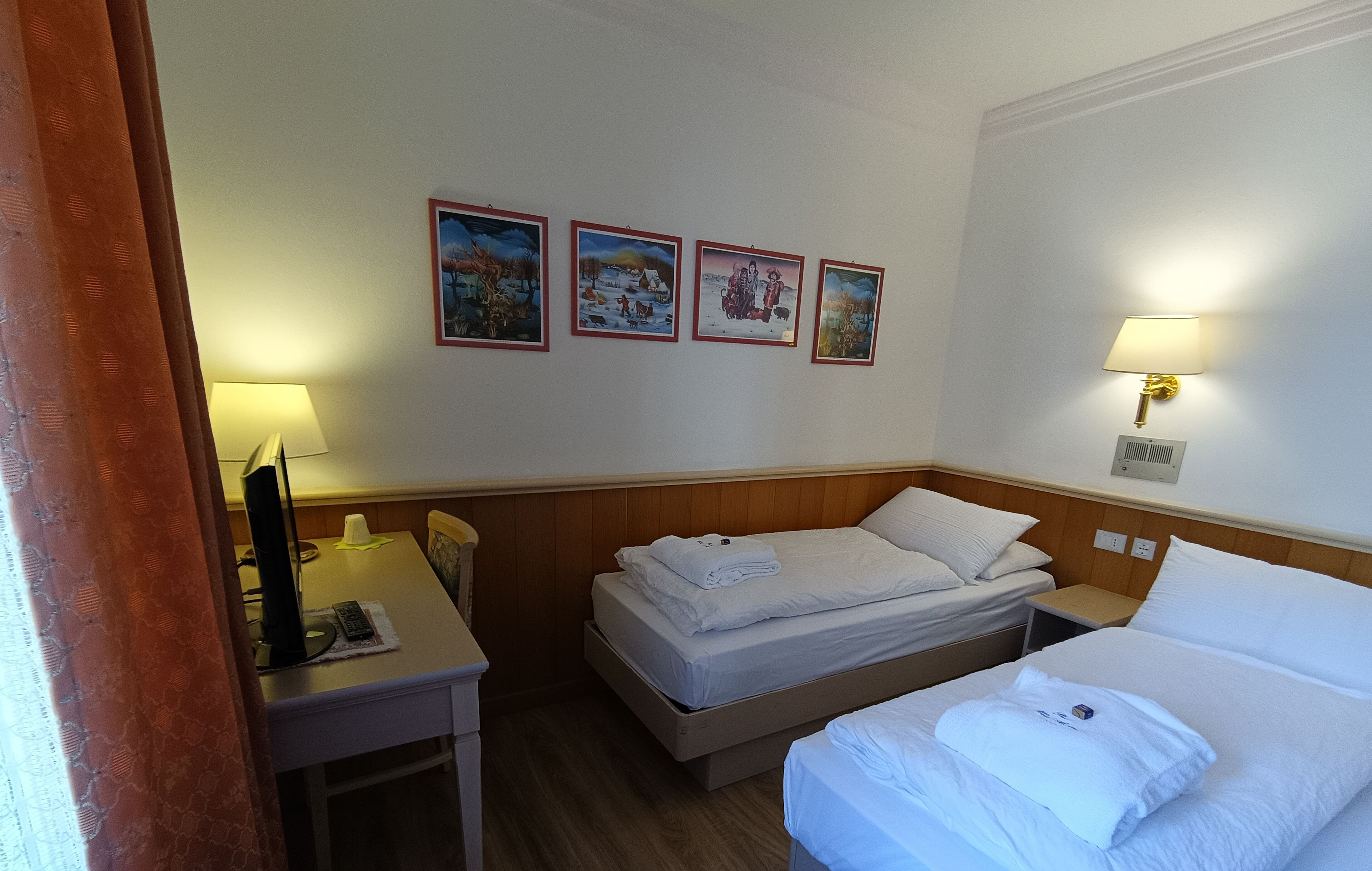 Une chambre d’hôtel avec deux lits, un bureau, une lampe et quatre tableaux encadrés au mur.