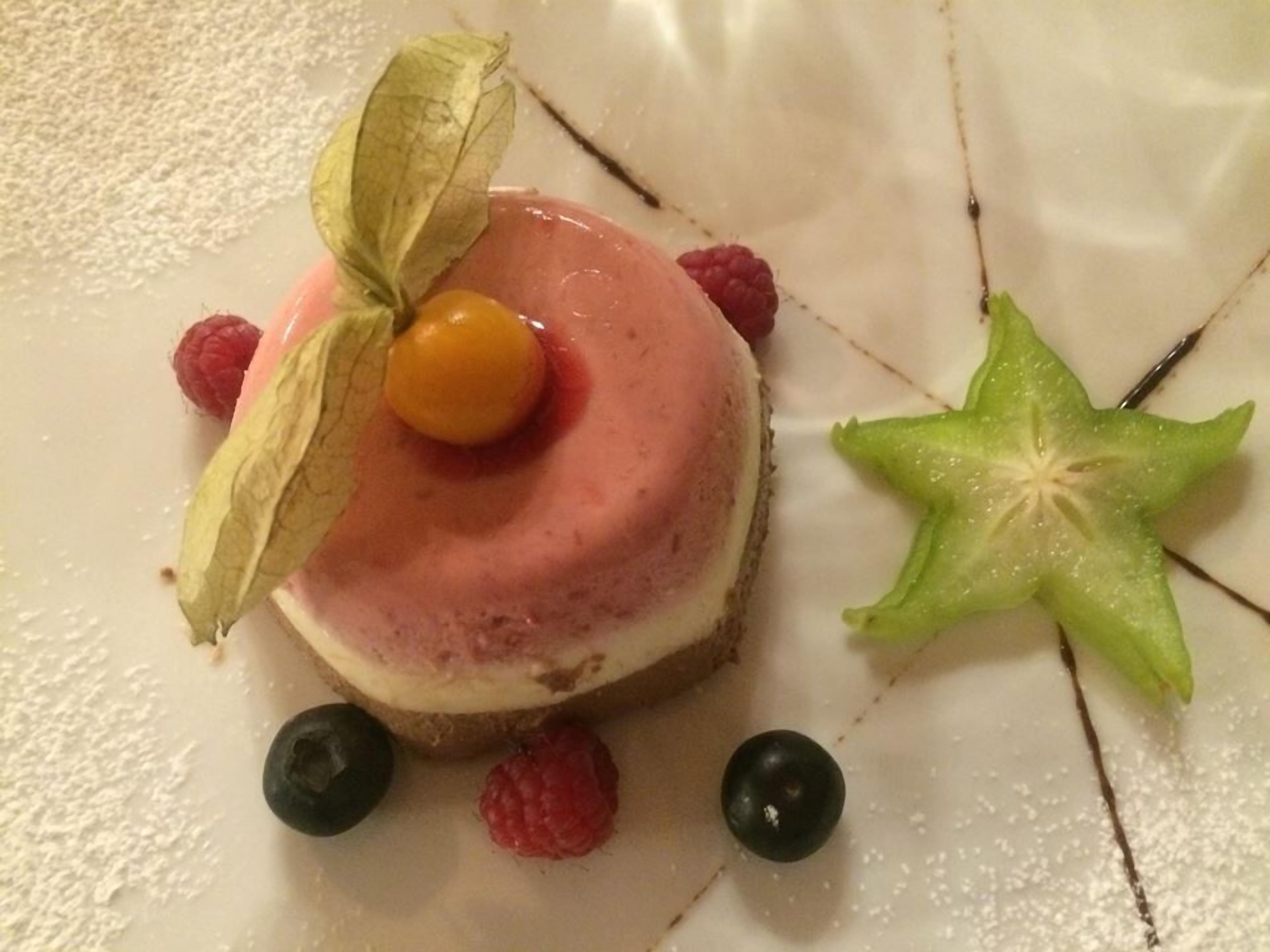Un dessert surmonté d’une cerise, entouré de framboises et de myrtilles, le tout présenté sur une assiette blanche.