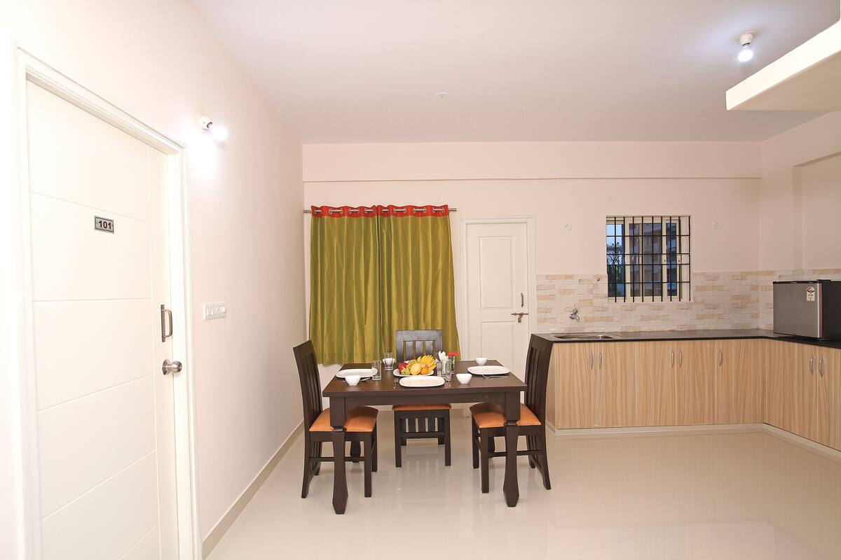Un comedor con una mesa dispuesta para cuatro, una cocina con refrigerador y una puerta con el número 101.