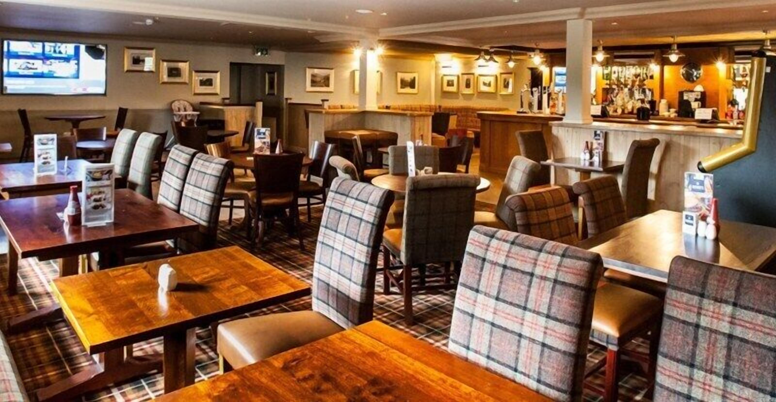 A restaurant with wooden tables and plaid chairs, a bar area, and framed pictures on the walls.