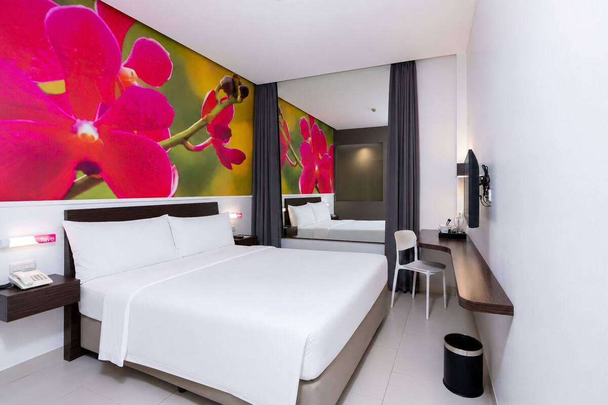 A hotel room with a large bed, a desk, a chair, a TV, and a wall with a floral mural.