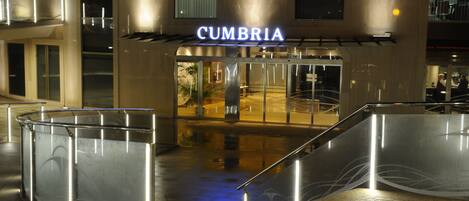 A modern hotel entrance with a prominent 'CUMBRIA' sign, a glass entryway, and a metal staircase leading to the lobby.