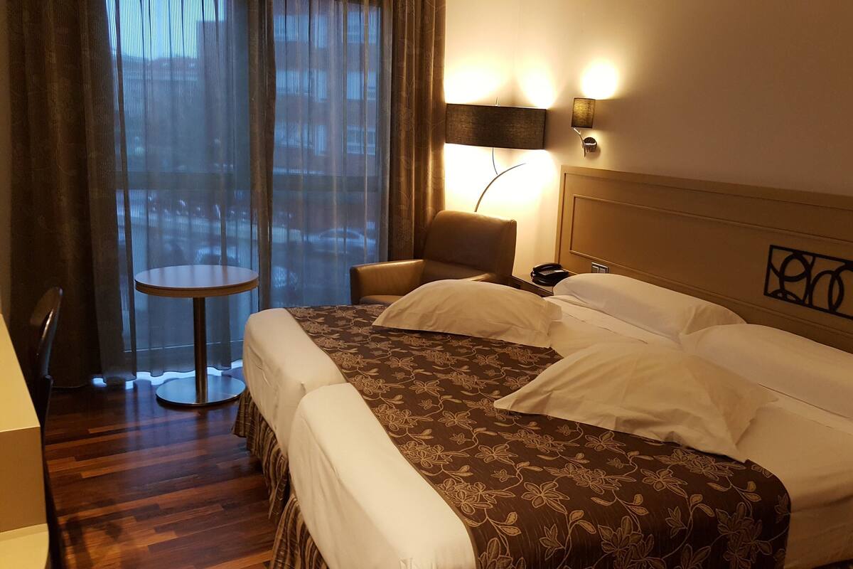 A hotel room with a large bed, a round table, a chair, and a view through sheer curtains.